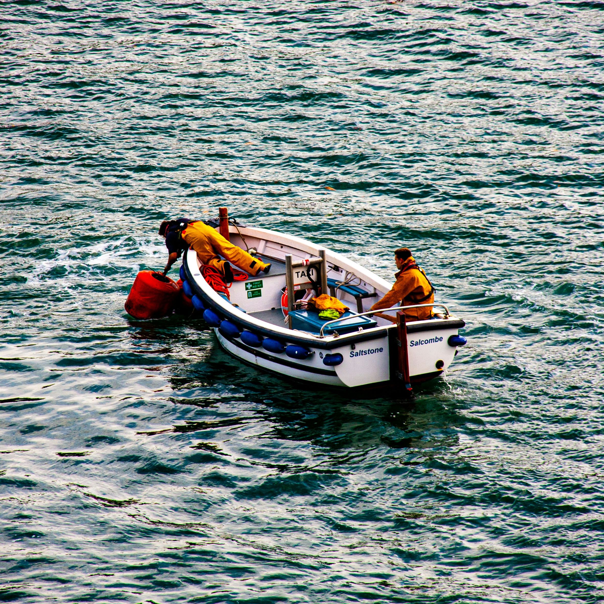 Two people in yellow raincoats work on a small white boat named Saltstone Salcombe on the open water.