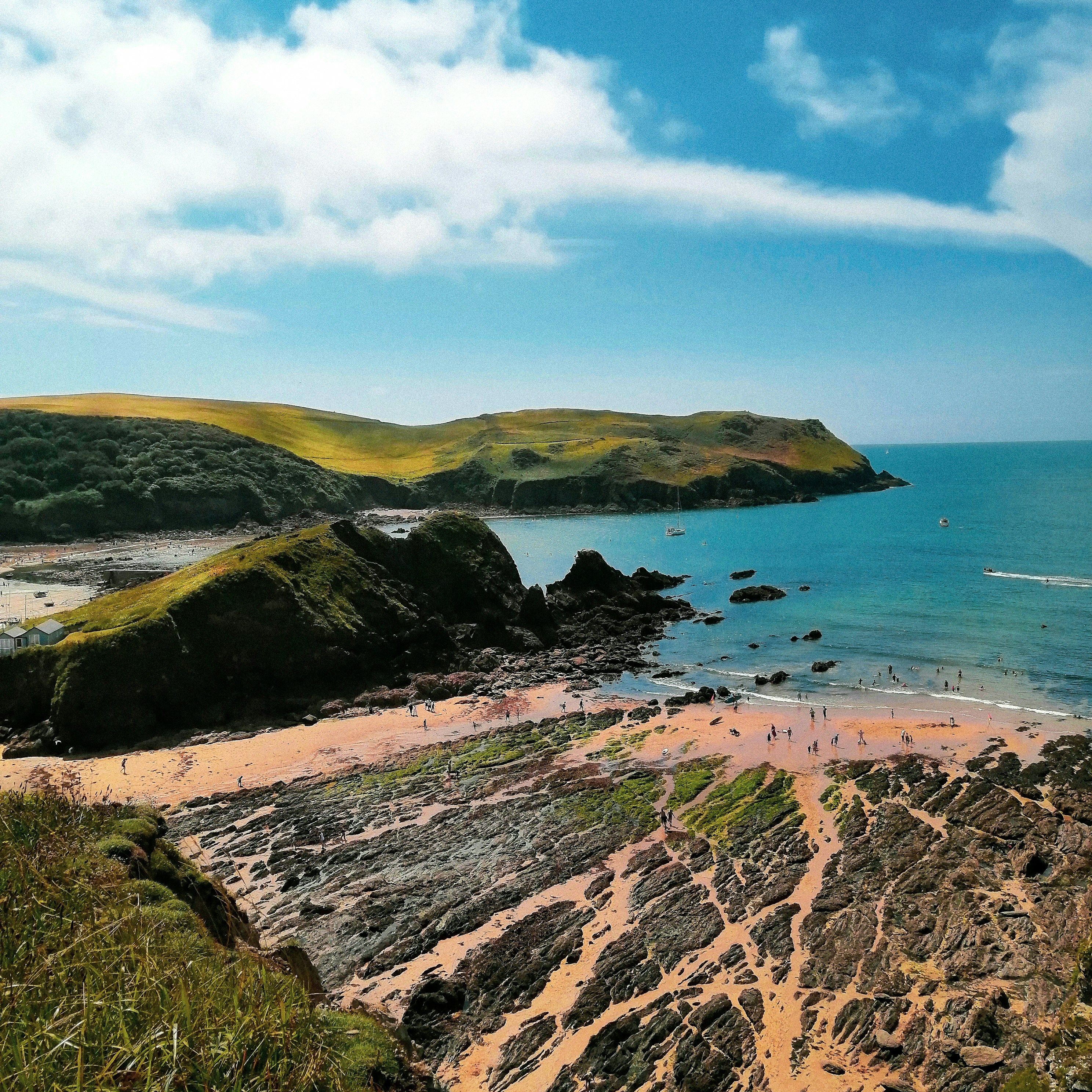 Coastal landscape with sandy beach, rocky formations, and grassy cliffs under a blue sky.