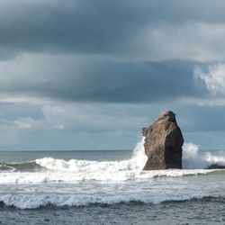 Large rock formation being hit by ocean waves under a cloudy sky