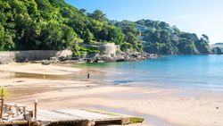A scenic beach with clear blue water, sandy shore, lush green trees, and stone walls in the background.