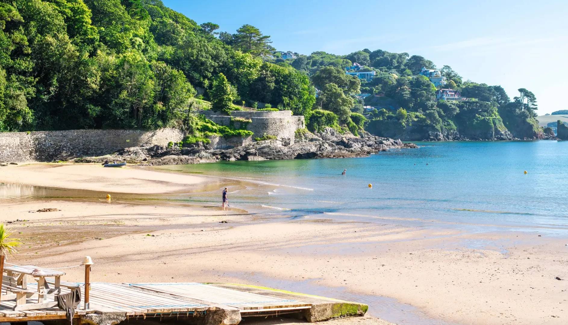 A scenic beach with clear blue water, sandy shore, lush green trees, and stone walls in the background.