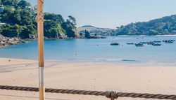 View of a sandy beach with boats floating on calm blue water, hills and houses in the background
