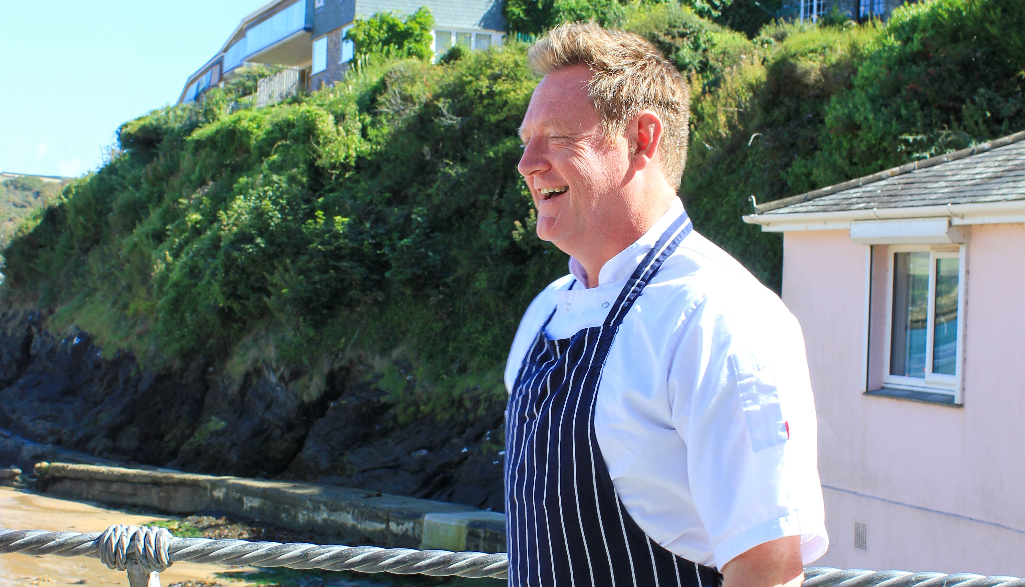 Chef wearing a striped apron standing outdoors near a beachside railing with houses and greenery in the background.