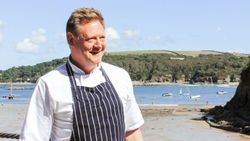 Chef wearing a striped apron standing on a sandy beach with boats and hills in the background.