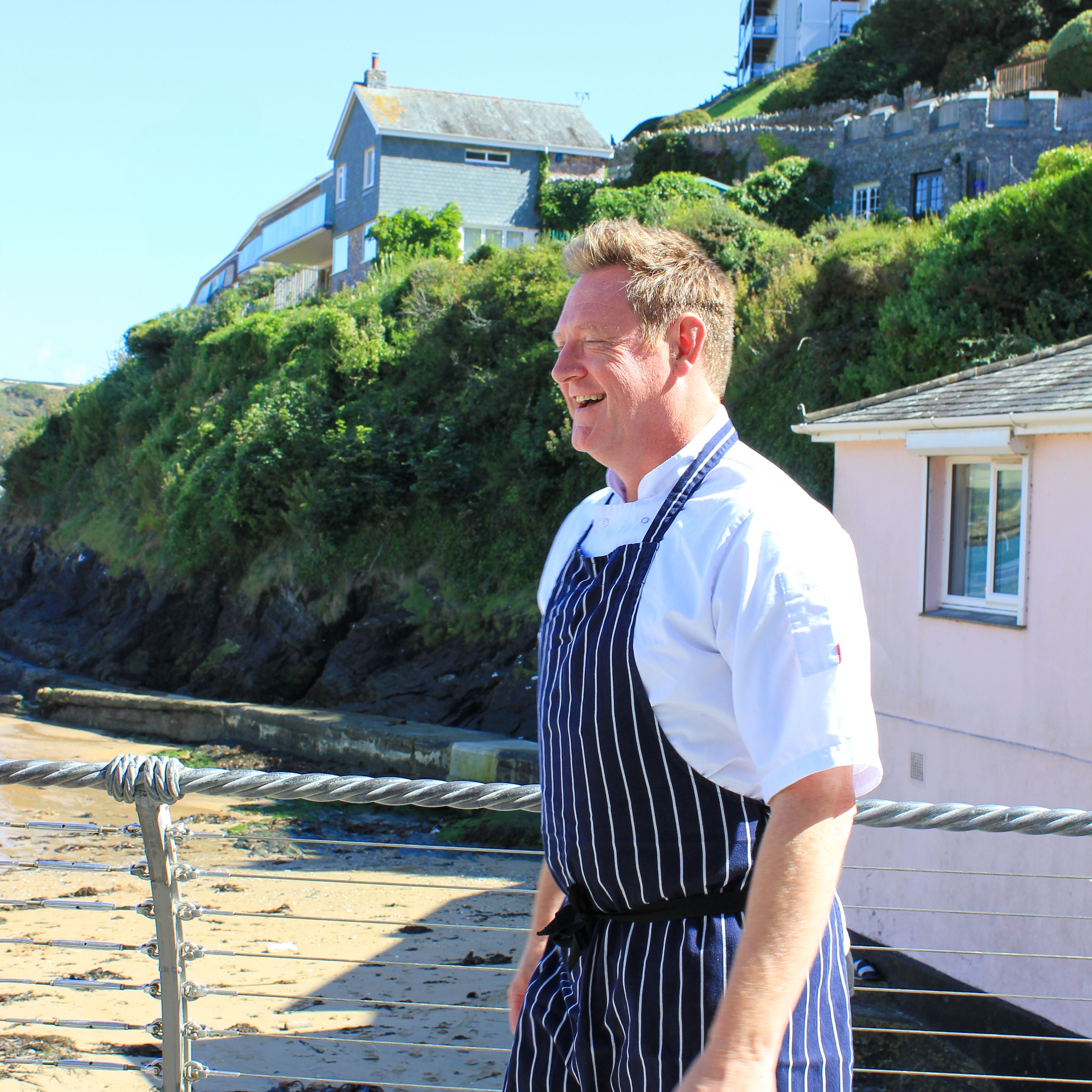 Chef wearing a striped apron standing outside near a beach and houses.