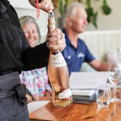 A server opening a bottle of rosé wine at a wooden table with two people seated in the background.