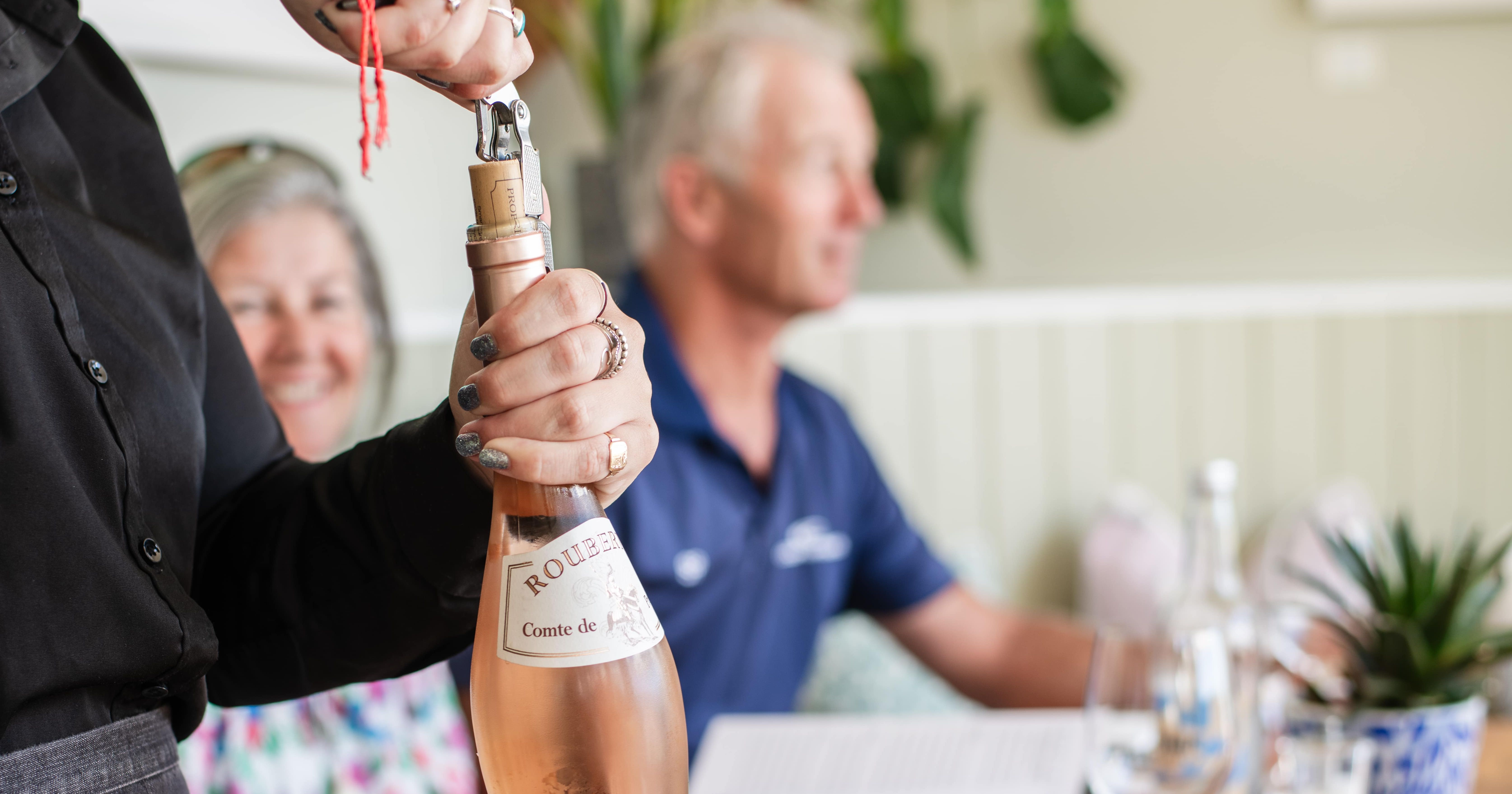 A server opening a bottle of rosé wine at a wooden table with two people seated in the background.
