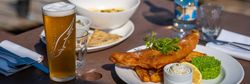 Plate of fish and chips with mushy peas, a lemon wedge, and tartar sauce, a glass of beer, a bottle of water, and other dishes on a table outdoors.