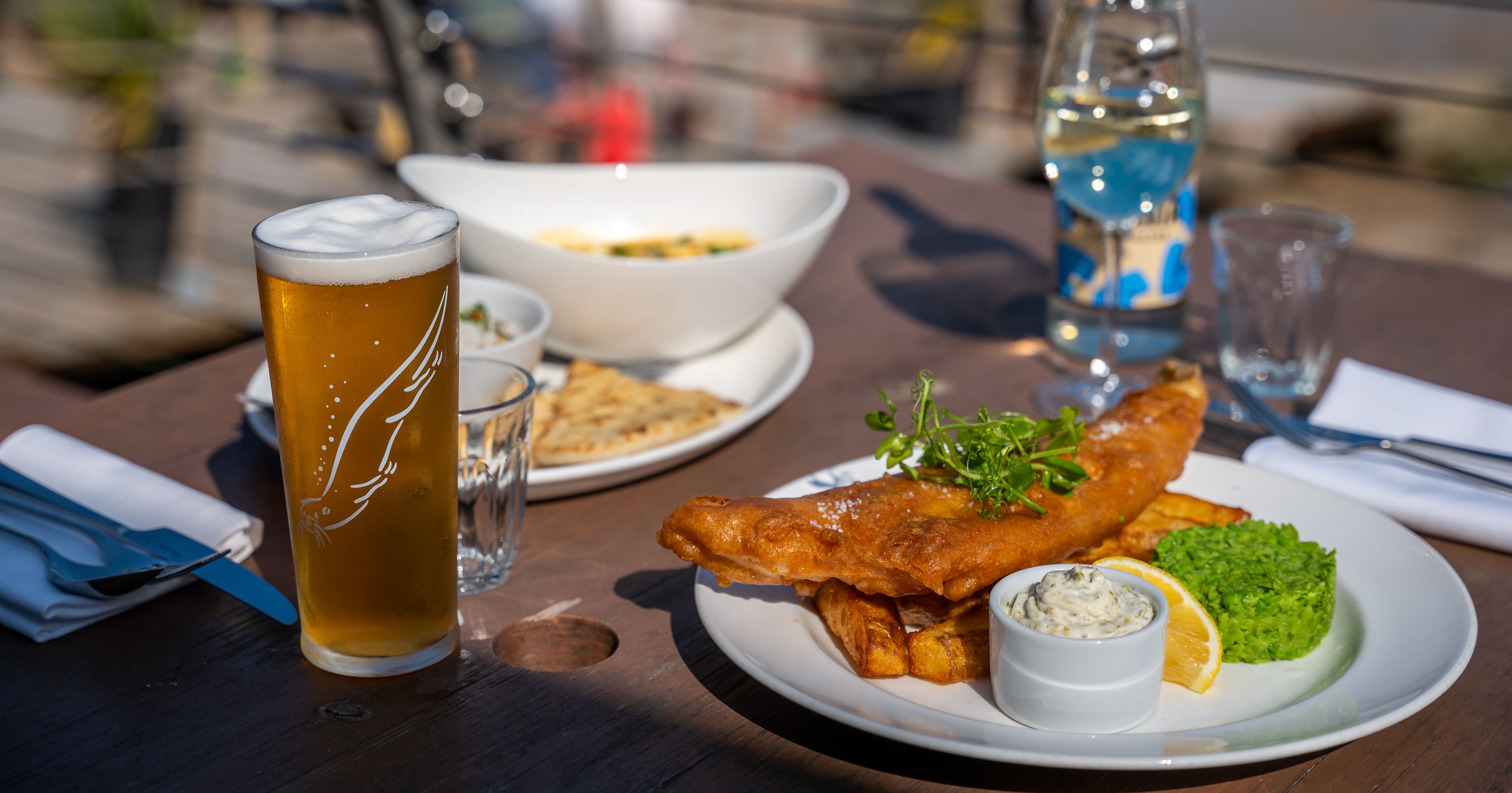 Plate of fish and chips with mushy peas, a lemon wedge, and tartar sauce, a glass of beer, a bottle of water, and other dishes on a table outdoors.