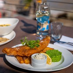 Plate of fish and chips with mushy peas, a lemon wedge, and tartar sauce, a glass of beer, a bottle of water, and other dishes on a table outdoors.