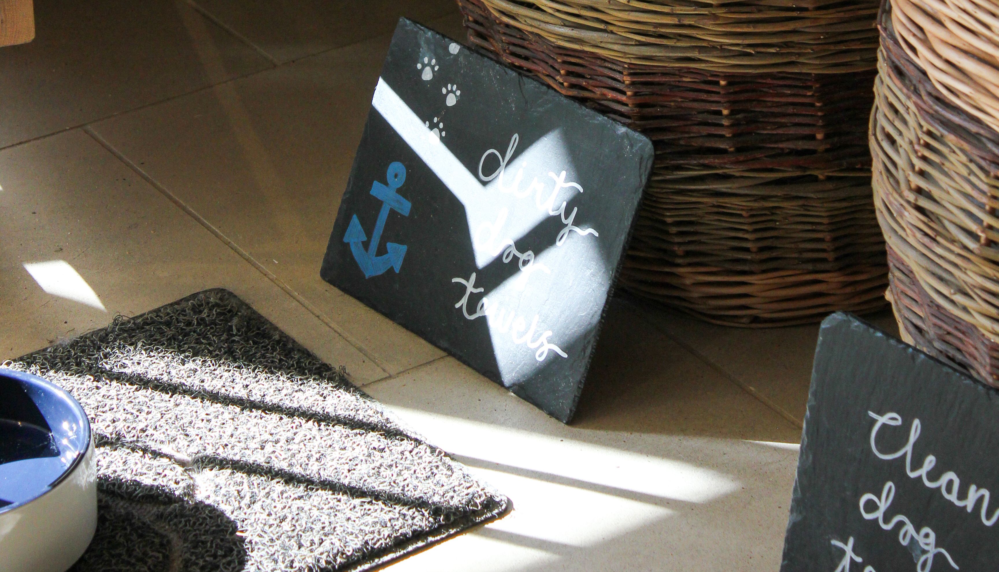 Wicker baskets under a staircase with labeled signs for dirty and clean dog towels, and a dog bowl on a mat.