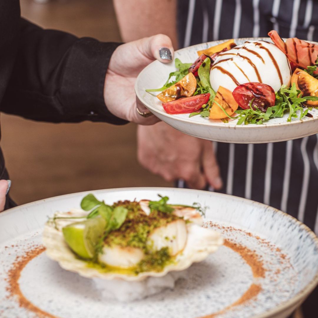 Two plates of gourmet summer dishes, one with a salad of mozzarella and colorful tomatoes, and another with scallops served in a shell with herbs and lime.