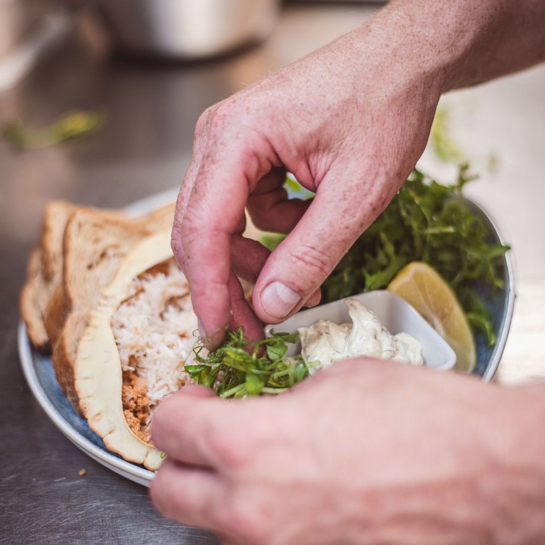 Hands garnishing a plate with fresh greens, pita bread, a wedge of lemon, and a small dish of creamy sauce.