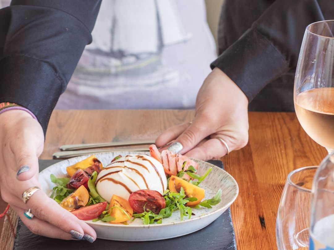 A person serving a fresh salad with burrata cheese, heirloom tomatoes, greens, and balsamic glaze on a wooden table.
