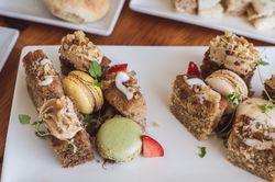 A plate of assorted pastries including macarons, cake slices with frosting and nuts, and garnished with herbs and strawberry pieces.