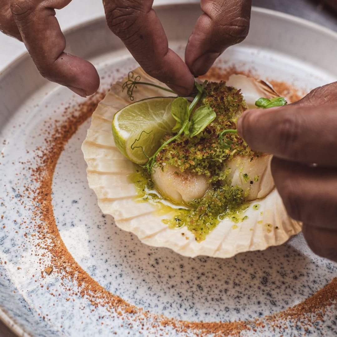 Chef garnishing scallop dish with herbs and lime in a shell on a decorative plate