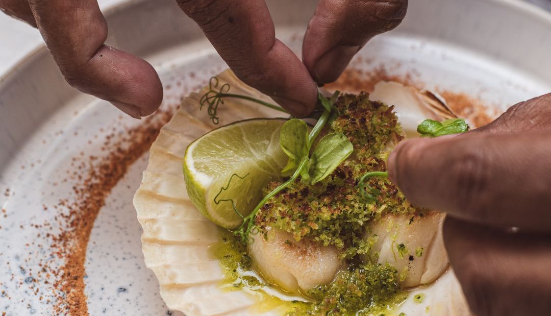 Chef garnishing scallop dish with herbs and lime in a shell on a decorative plate