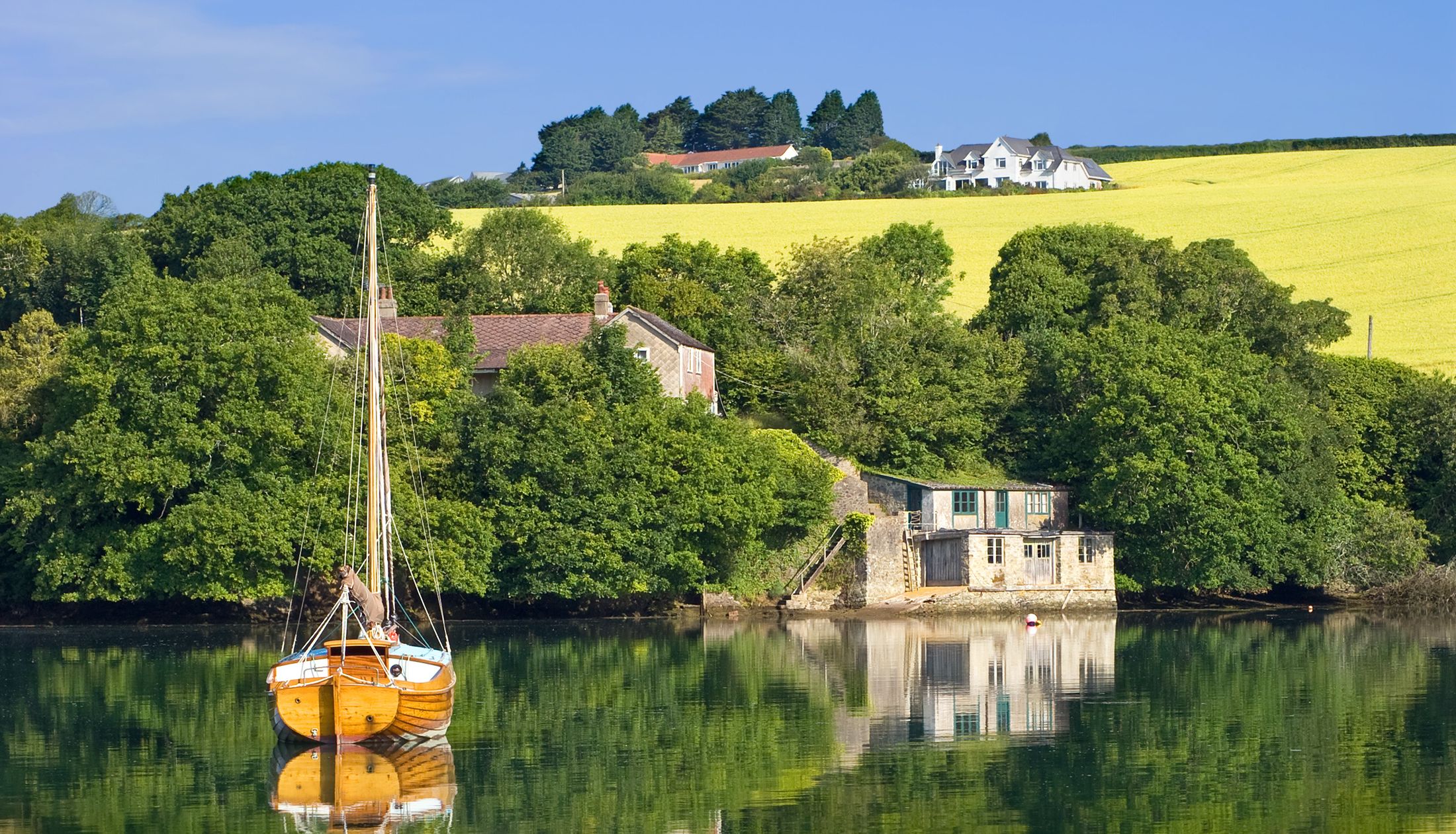 Wooden sailboat on calm water with farmhouse and yellow field in the background