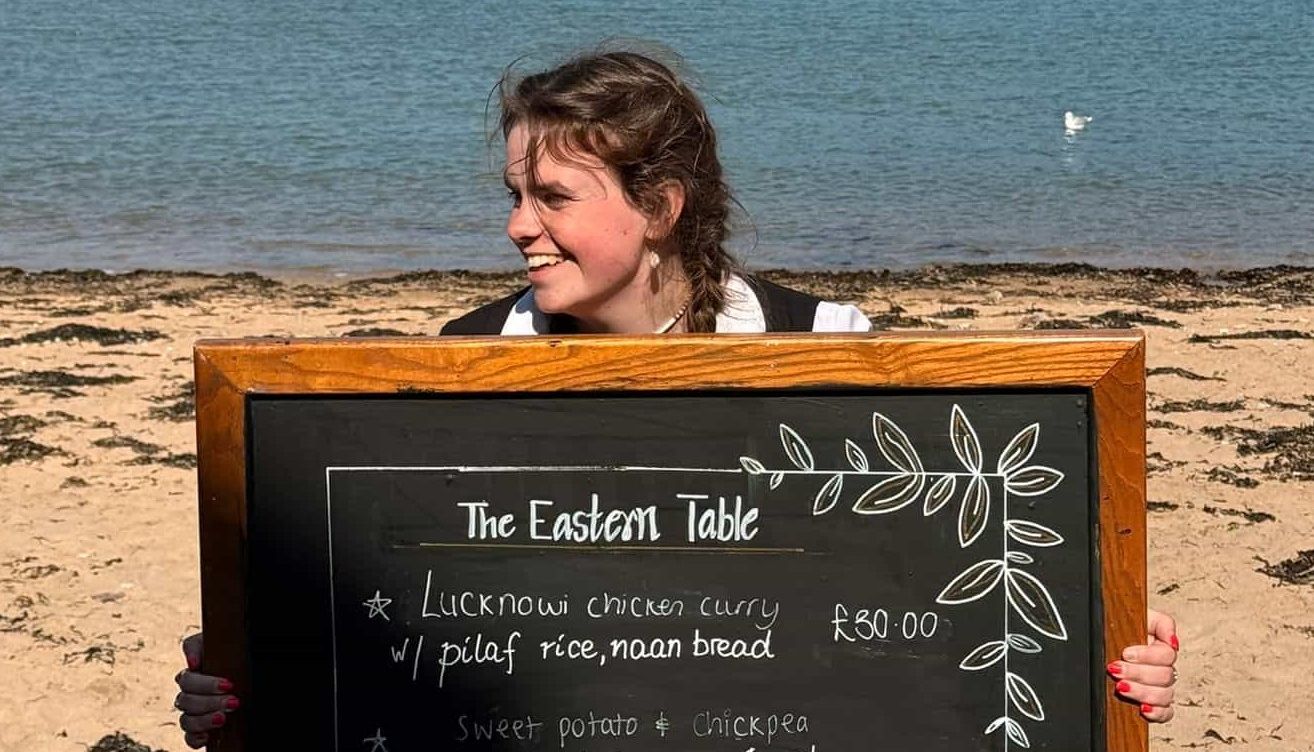 Woman holding a chalkboard menu on a sandy beach with the sea and hills in the background.