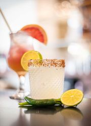 Close-up of a cocktail in a textured glass with a chili pepper and lime slice, with another cocktail blurred in the background.