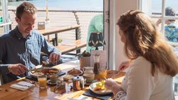 A man and woman enjoying breakfast at a table by a window overlooking a beach.