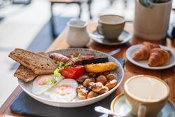 Full English breakfast with eggs, toast, sausage, mushrooms, tomato, black pudding, hash brown, and croissants on a table with coffee.