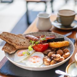 Full English breakfast with eggs, toast, sausage, mushrooms, tomato, black pudding, hash brown, and croissants on a table with coffee.