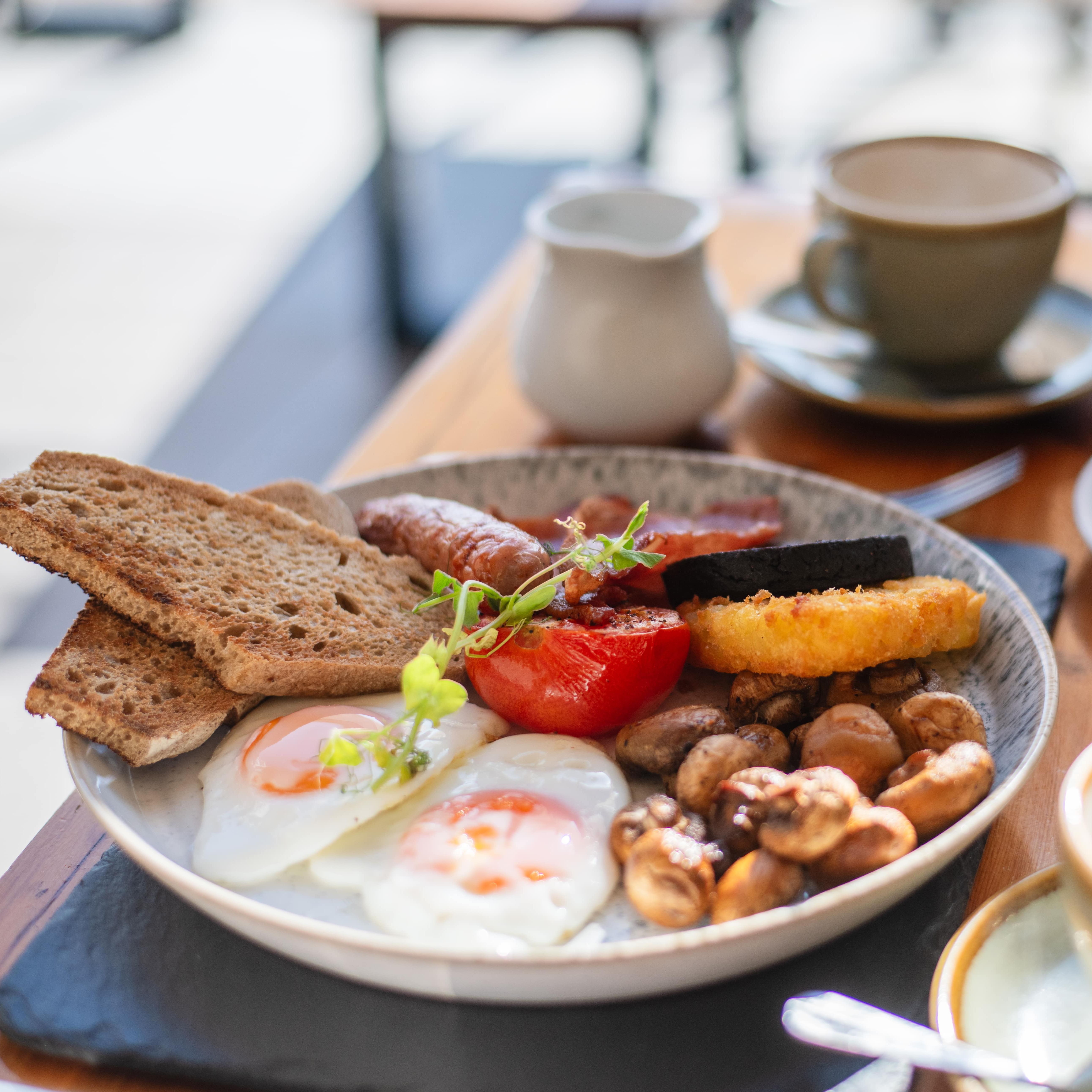 Full English breakfast with eggs, toast, sausage, mushrooms, tomato, black pudding, hash brown, and croissants on a table with coffee.