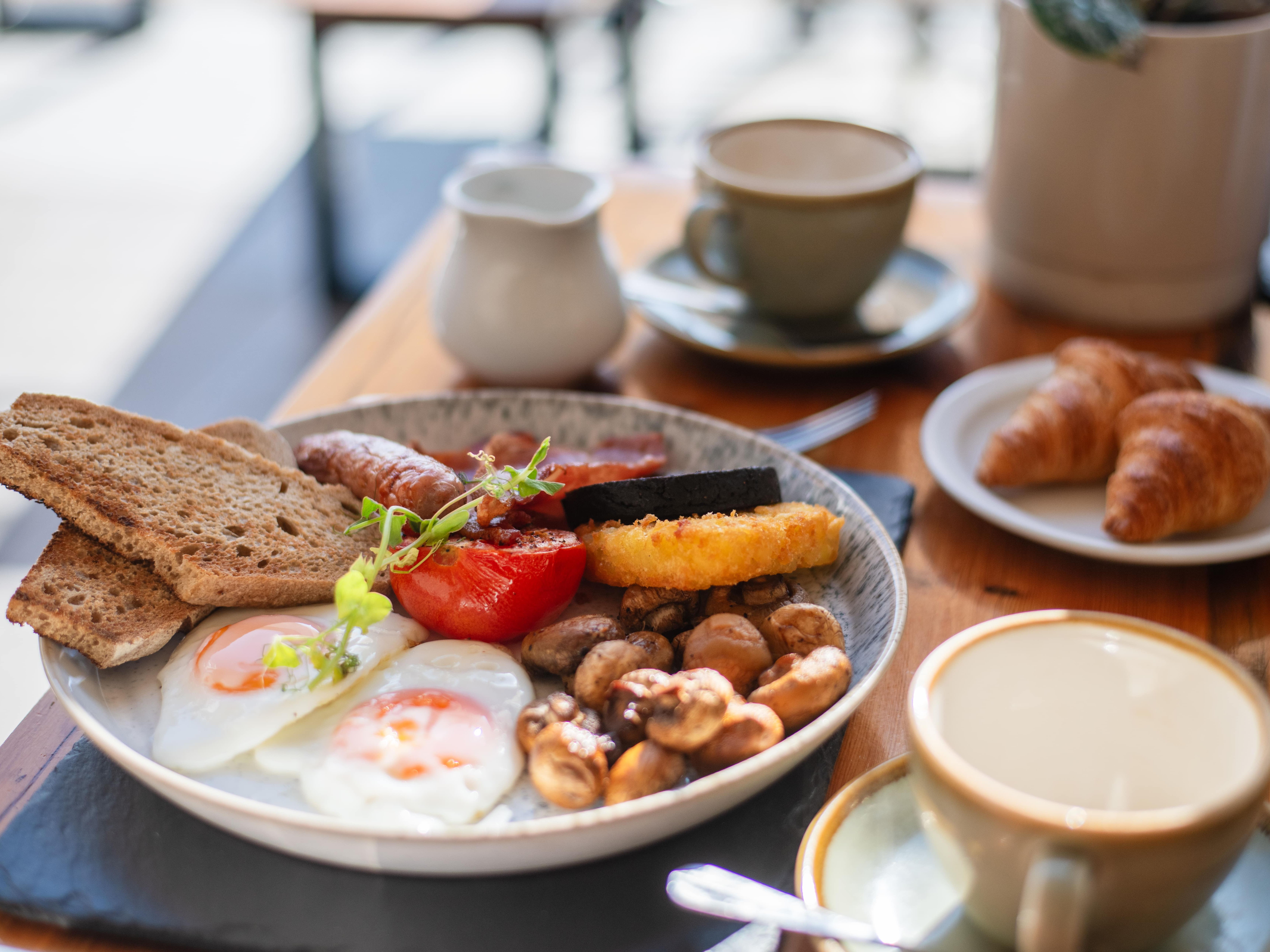 Full English breakfast with eggs, toast, sausage, mushrooms, tomato, black pudding, hash brown, and croissants on a table with coffee.