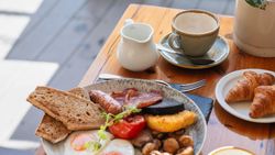 Full English breakfast with eggs, toast, sausage, bacon, and sides on a table with coffee cups and croissants.