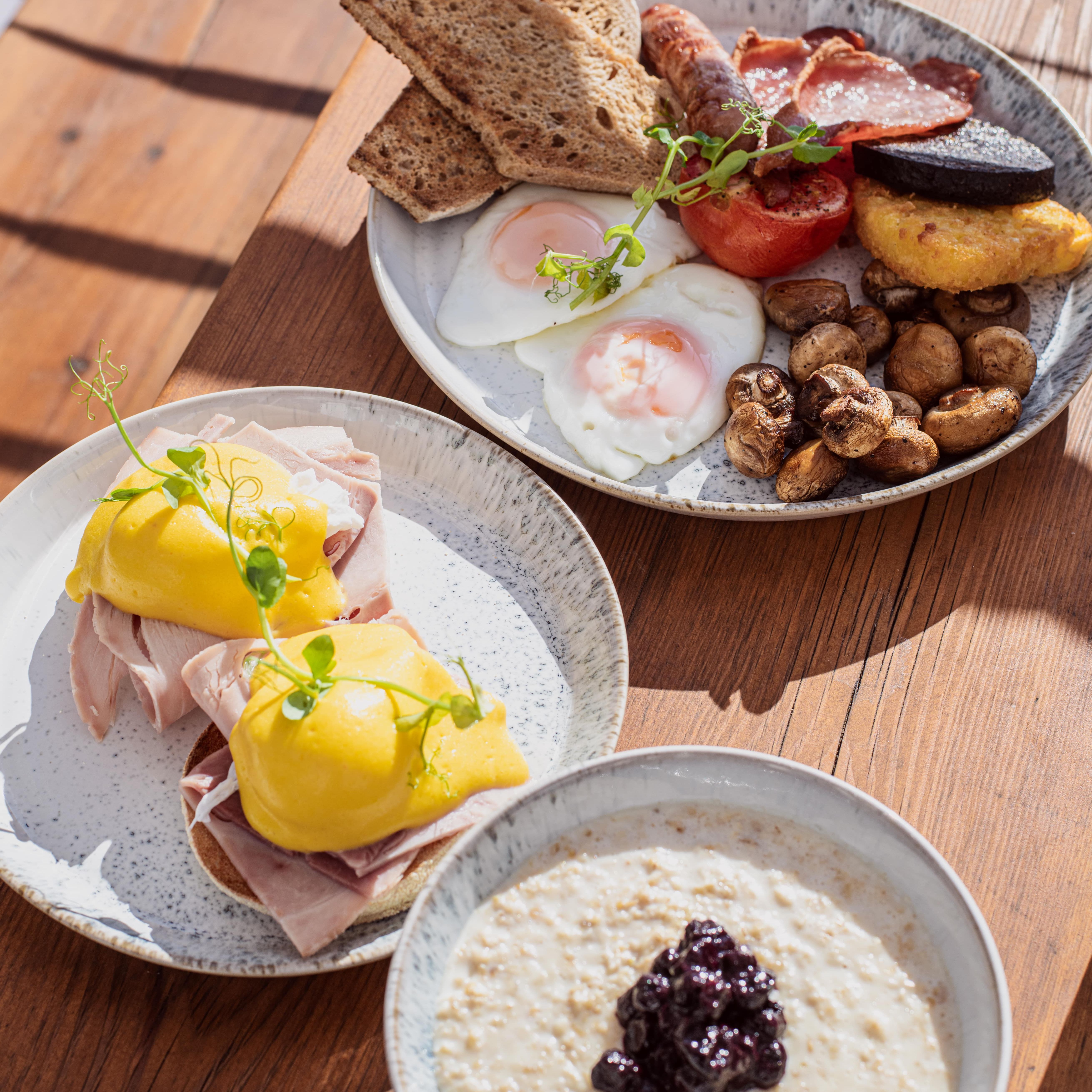 Three plates of breakfast food including eggs benedict, a traditional English breakfast, and a bowl of porridge with fruit compote.
