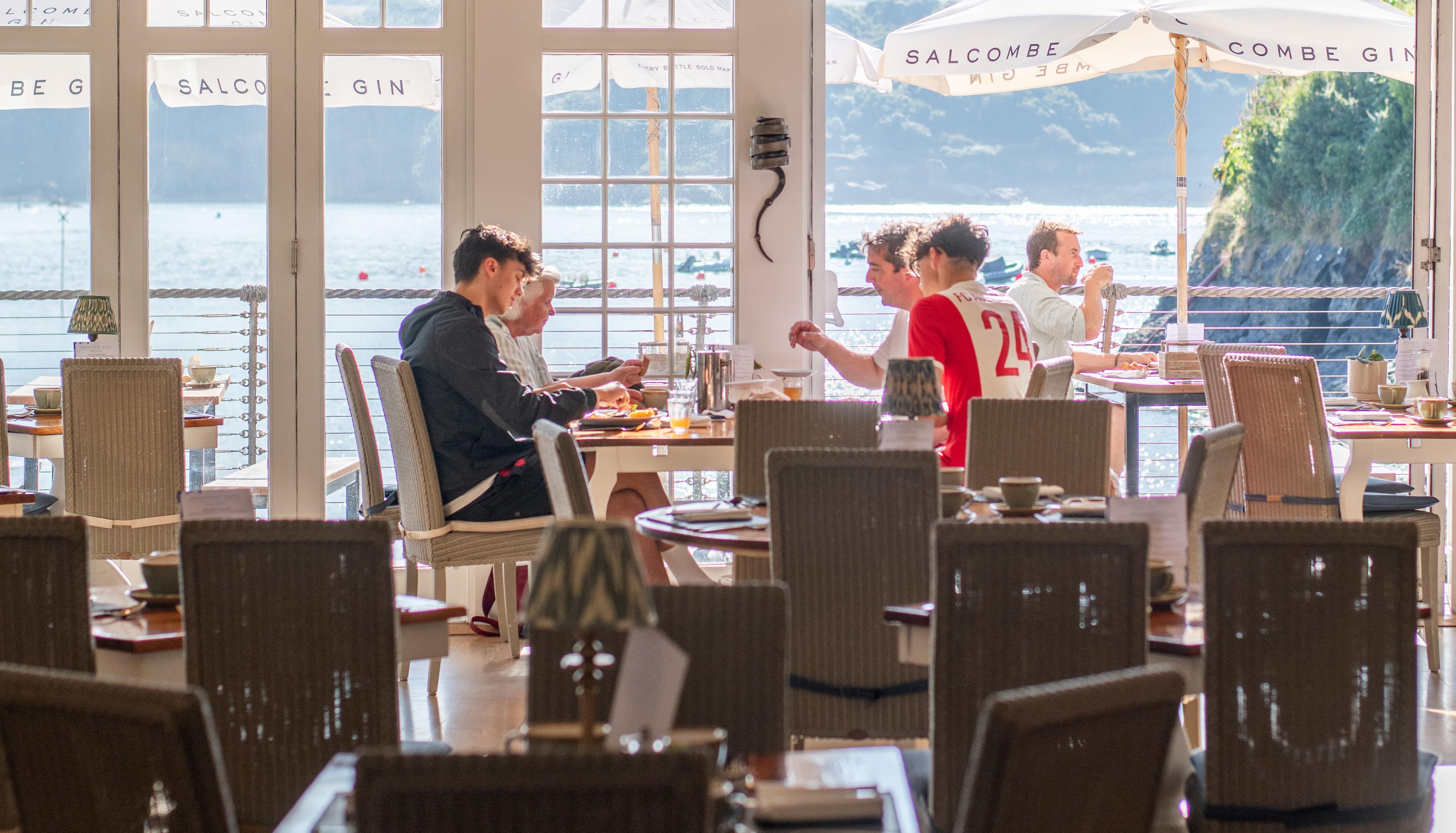 People dining inside a restaurant with large windows overlooking a waterfront view.
