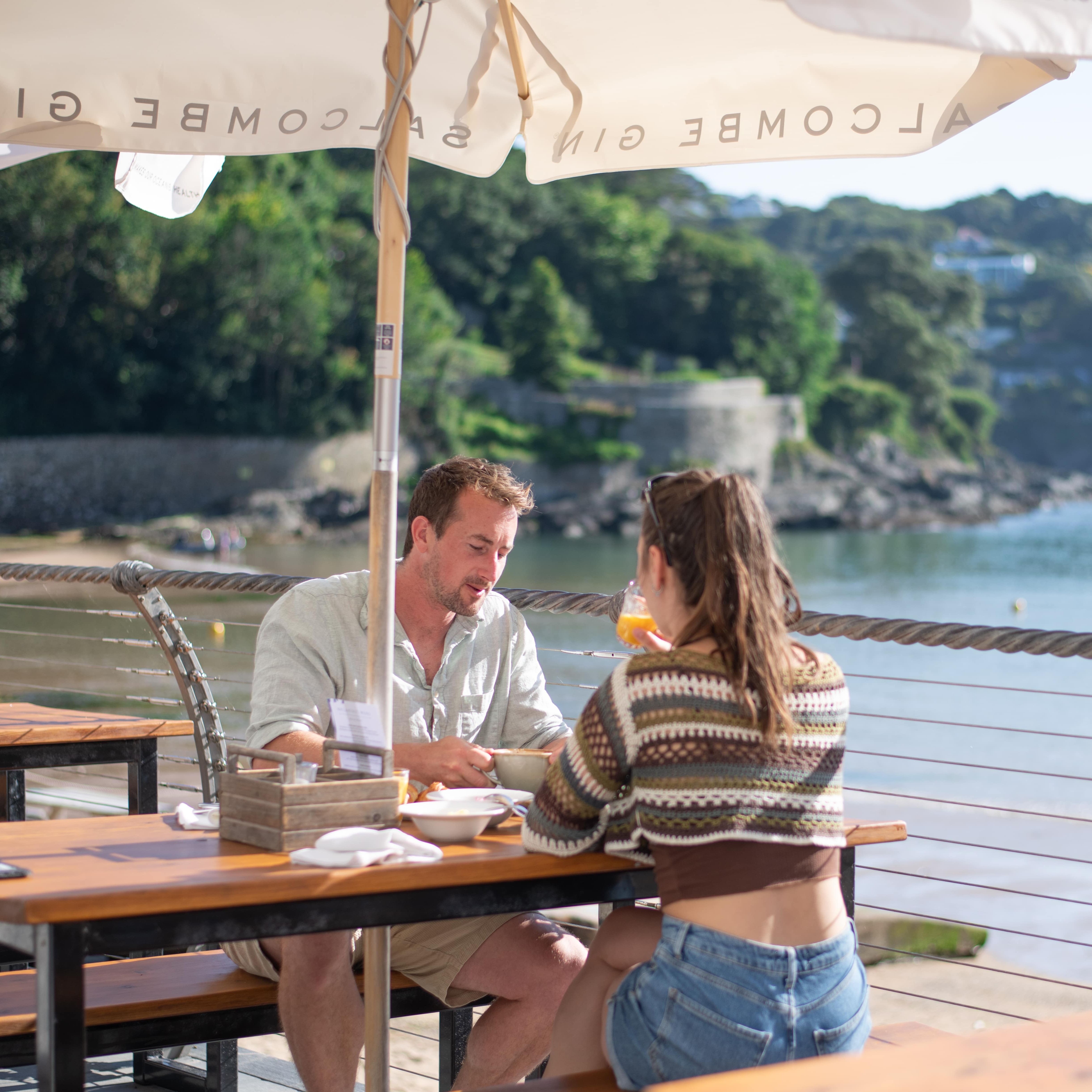 Two people dining at an outdoor restaurant by the water with trees and hills in the background.