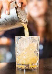 Bartender pouring a yellow cocktail over ice from a shaker into a glass