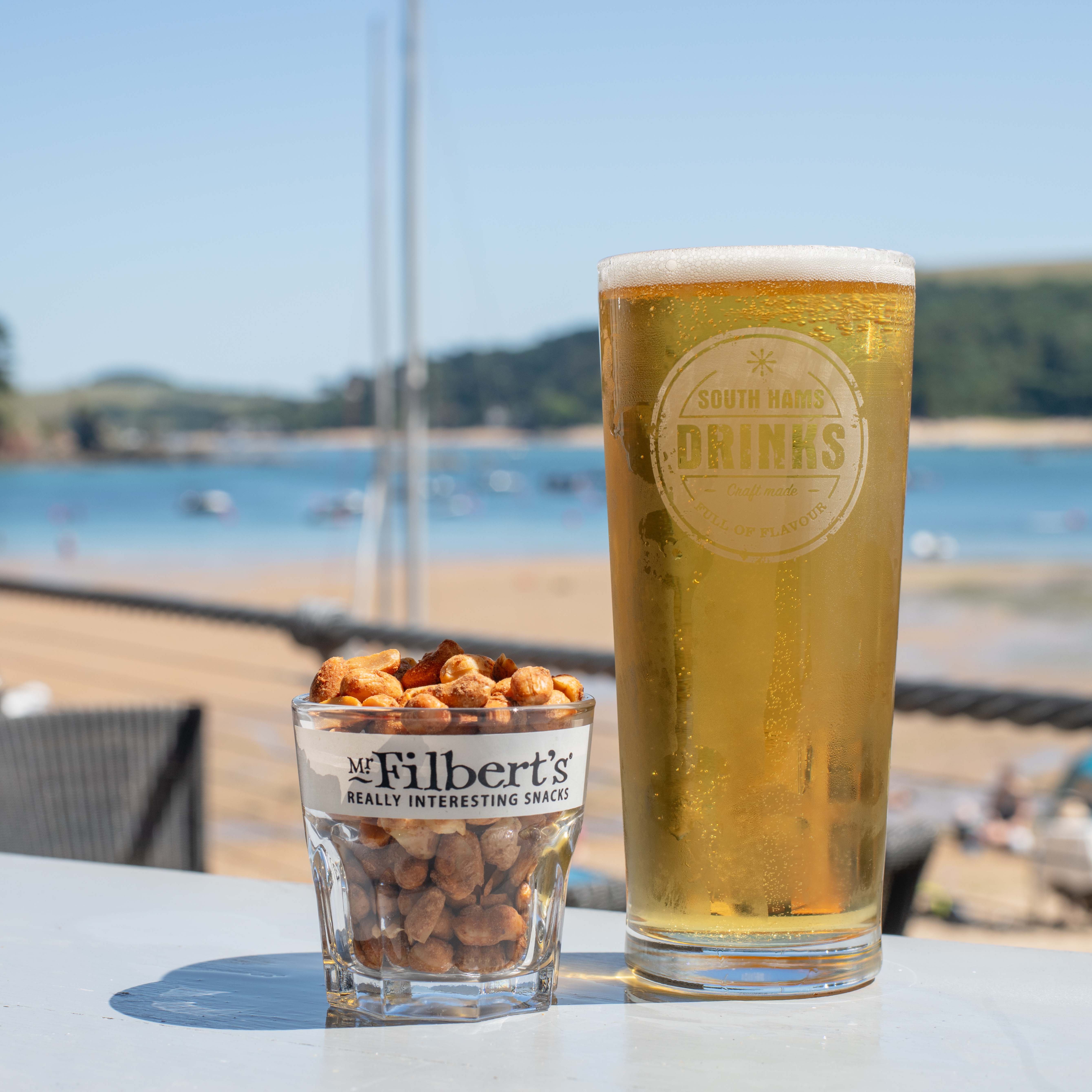 A glass of beer and a glass filled with mixed nuts on an outdoor table overlooking a beach.