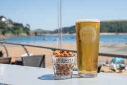 A glass of beer and a glass filled with mixed nuts on an outdoor table overlooking a beach.