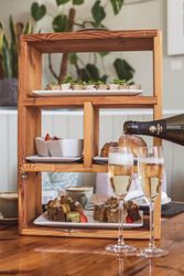 Wooden afternoon tea stand with a variety of sandwiches, scones, cakes, and two glasses of sparkling wine being poured.