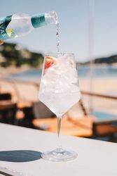 Sparkling water being poured into a wine glass filled with ice and a slice of citrus fruit on an outdoor table by the beach.