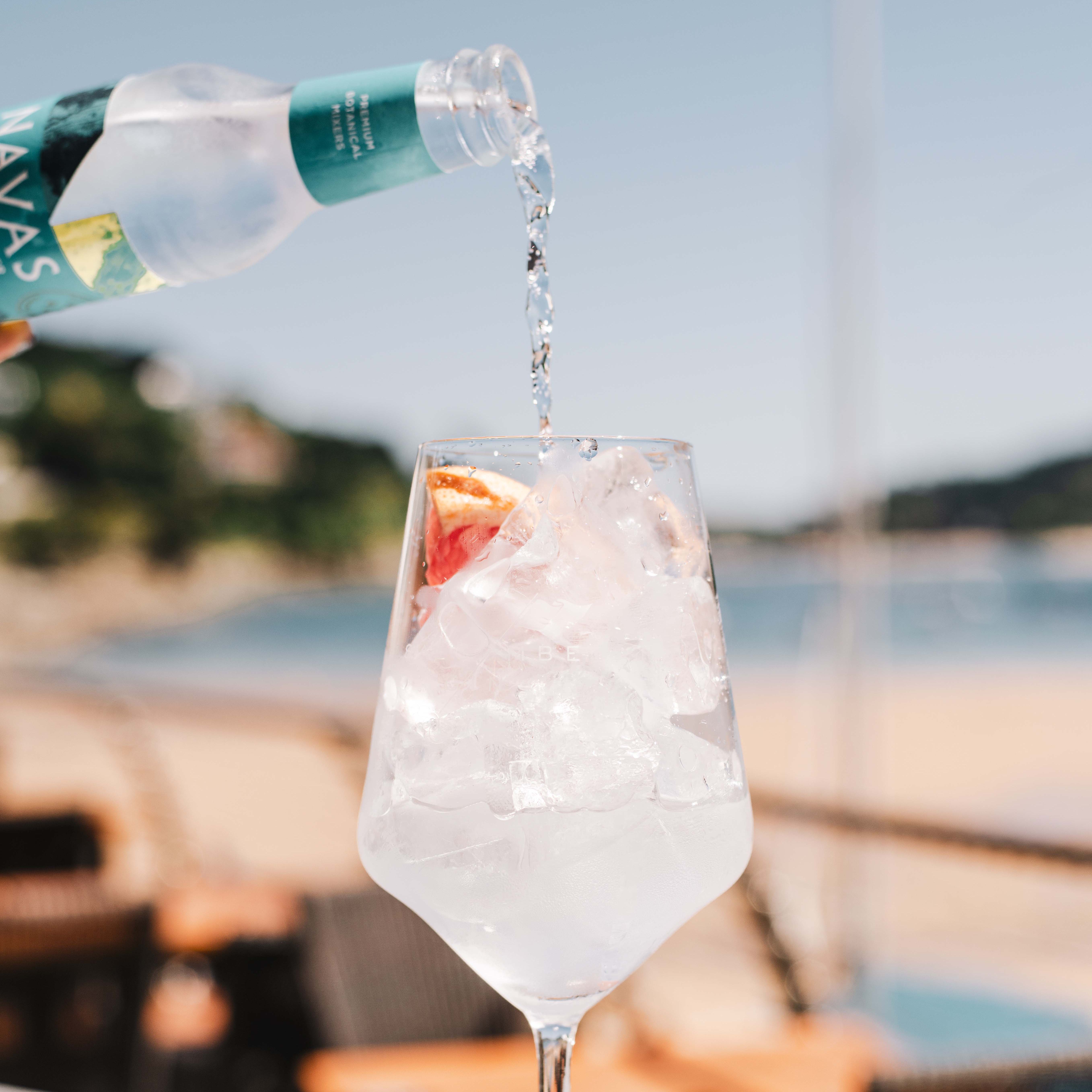 Sparkling water being poured into a wine glass filled with ice and a slice of citrus fruit on an outdoor table by the beach.