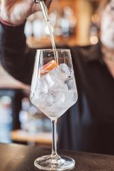 A person pouring a clear liquid into a wine glass filled with ice and a slice of citrus fruit.