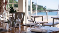 Elegant restaurant table set with glasses, napkins, and a plant, overlooking a sunny beach view through open doors.