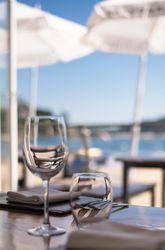 Wine glasses and napkins on a table at an outdoor restaurant with a blurry view of umbrellas and water in the background.