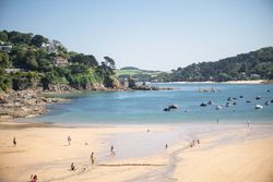 People walking and playing on a sandy beach with boats floating on calm blue water and green hills in the background.