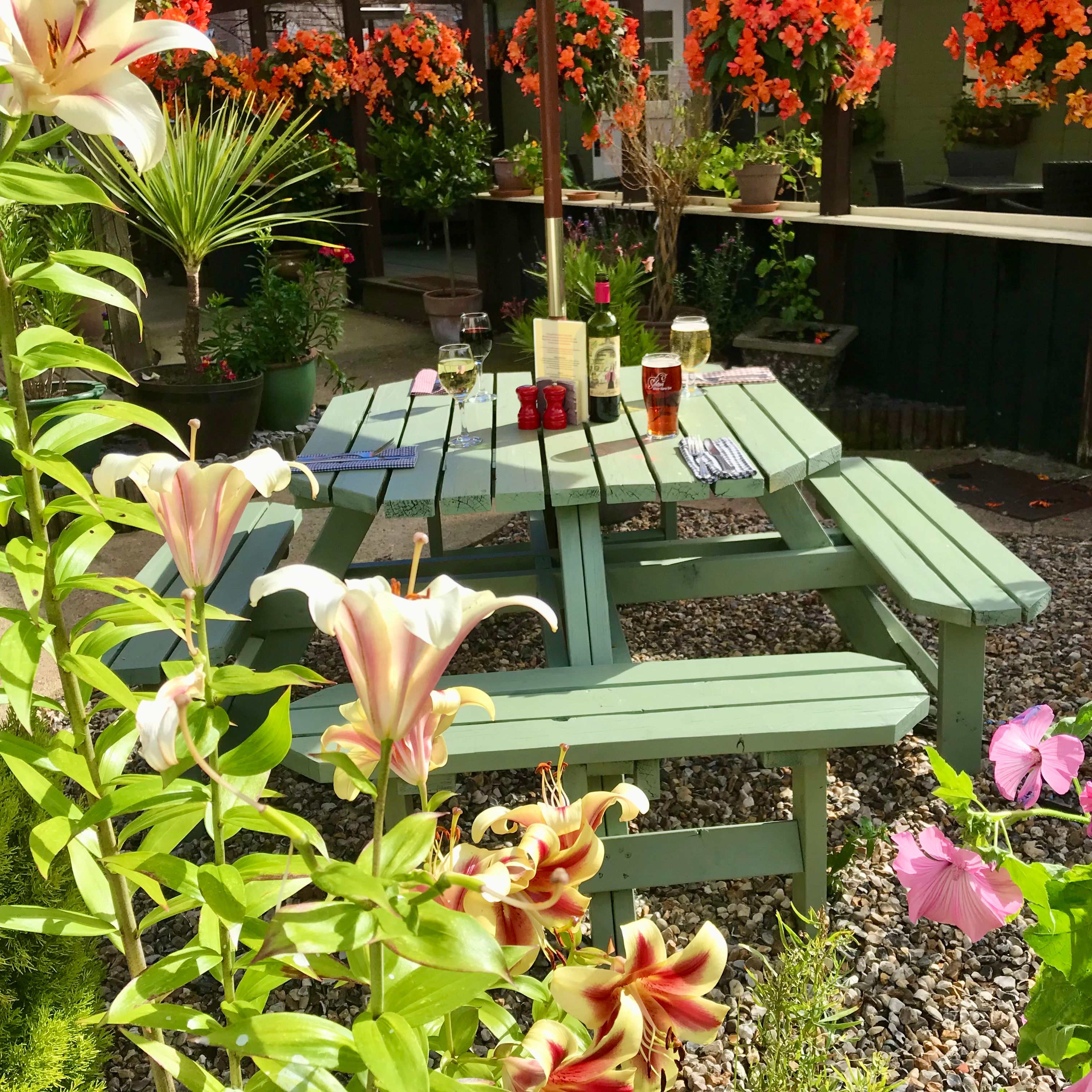 Outdoor garden seating area with green wooden picnic table, drinks on the table, and colorful flowers all around.