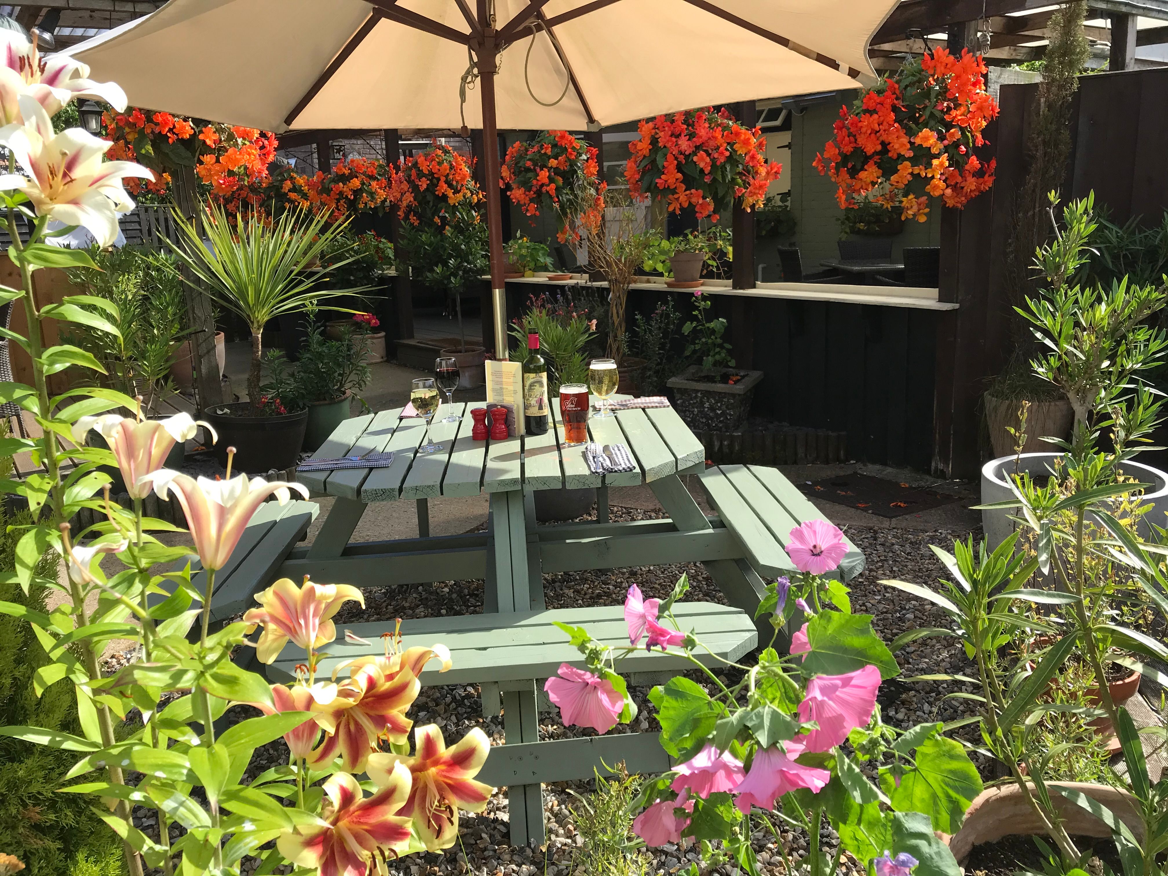 Sunny outdoor garden patio with green picnic table, vibrant flowers, drinks, and large umbrella