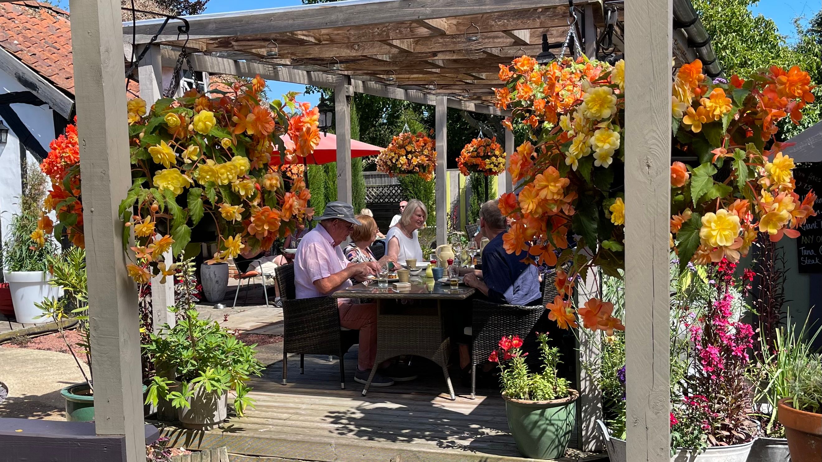 People enjoying a meal at an outdoor patio surrounded by vibrant hanging flower baskets.