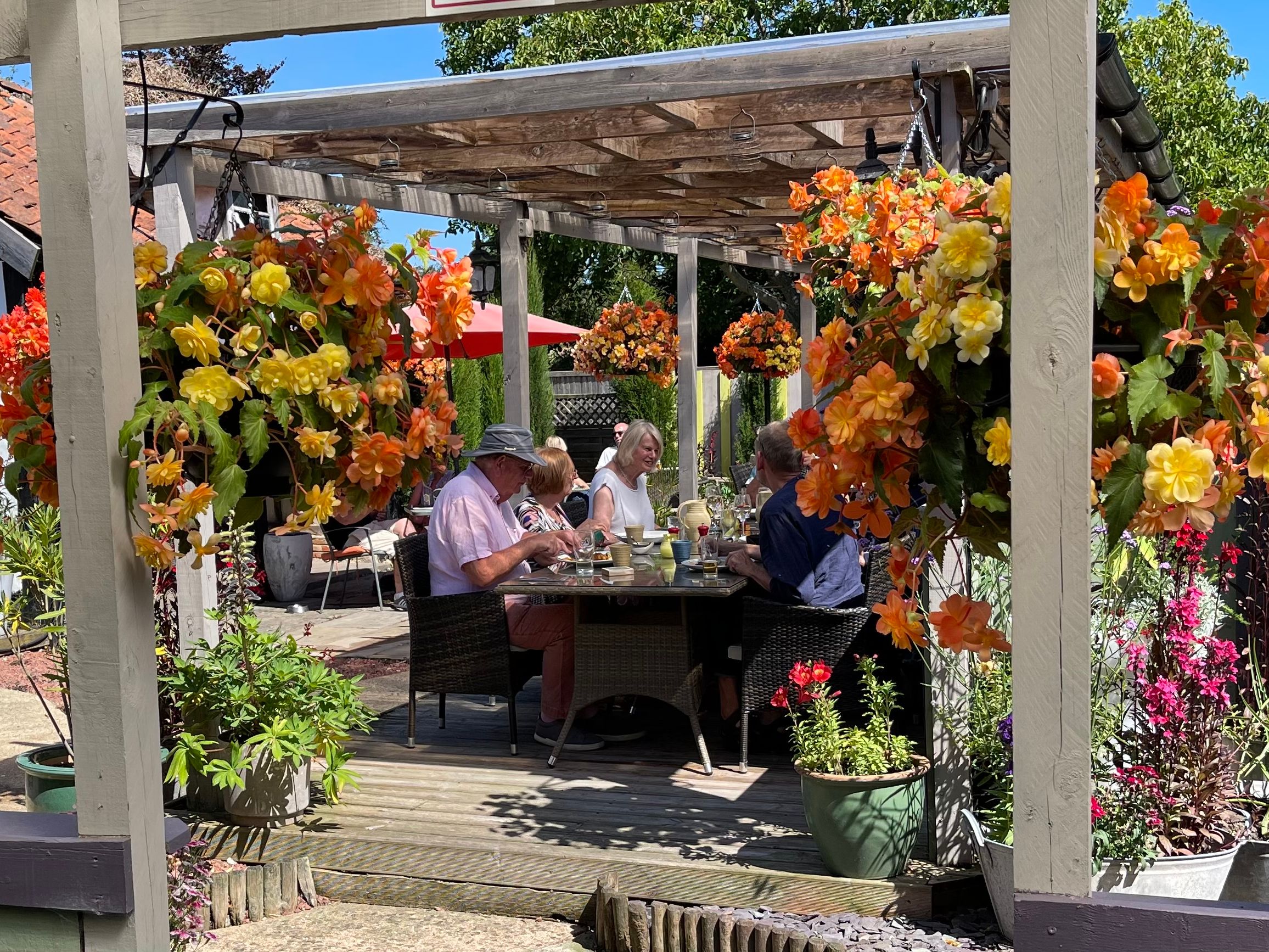 People enjoying a meal at an outdoor patio surrounded by vibrant hanging flower baskets.