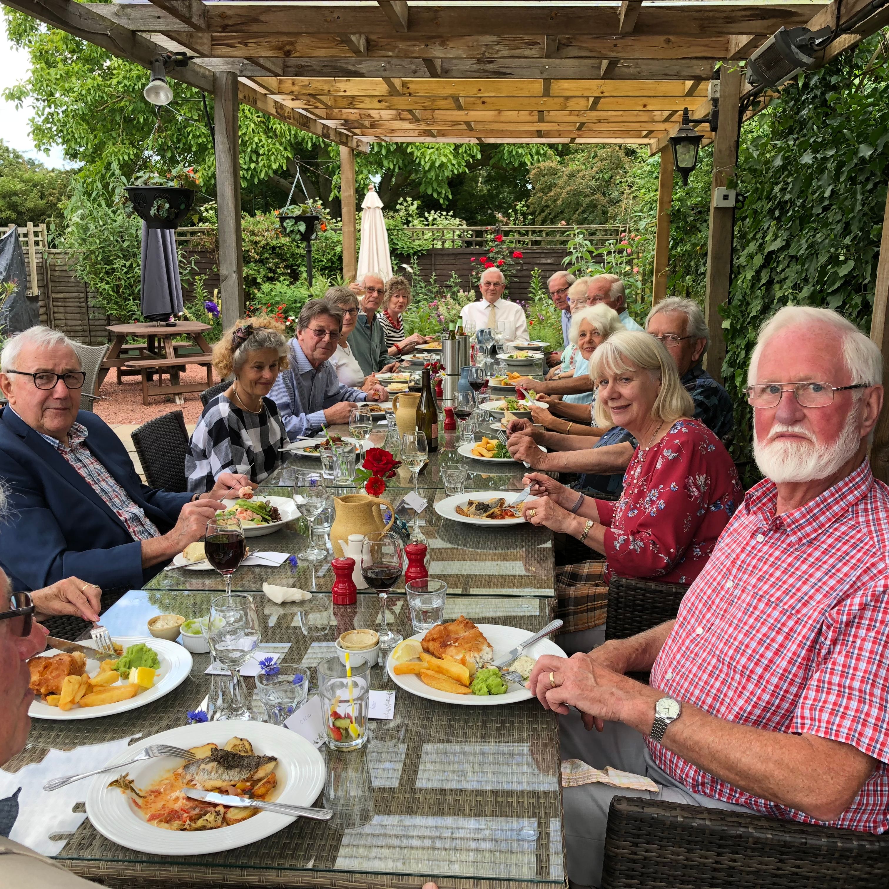 A group of elderly people enjoying a meal at a long outdoor dining table under a wooden pergola.