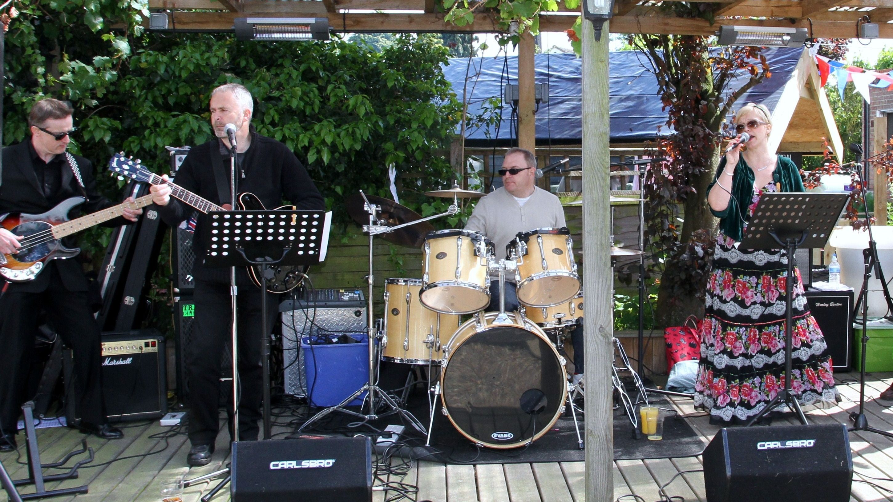 A band performs outdoors under a wooden pergola decorated with red, white, and blue bunting.