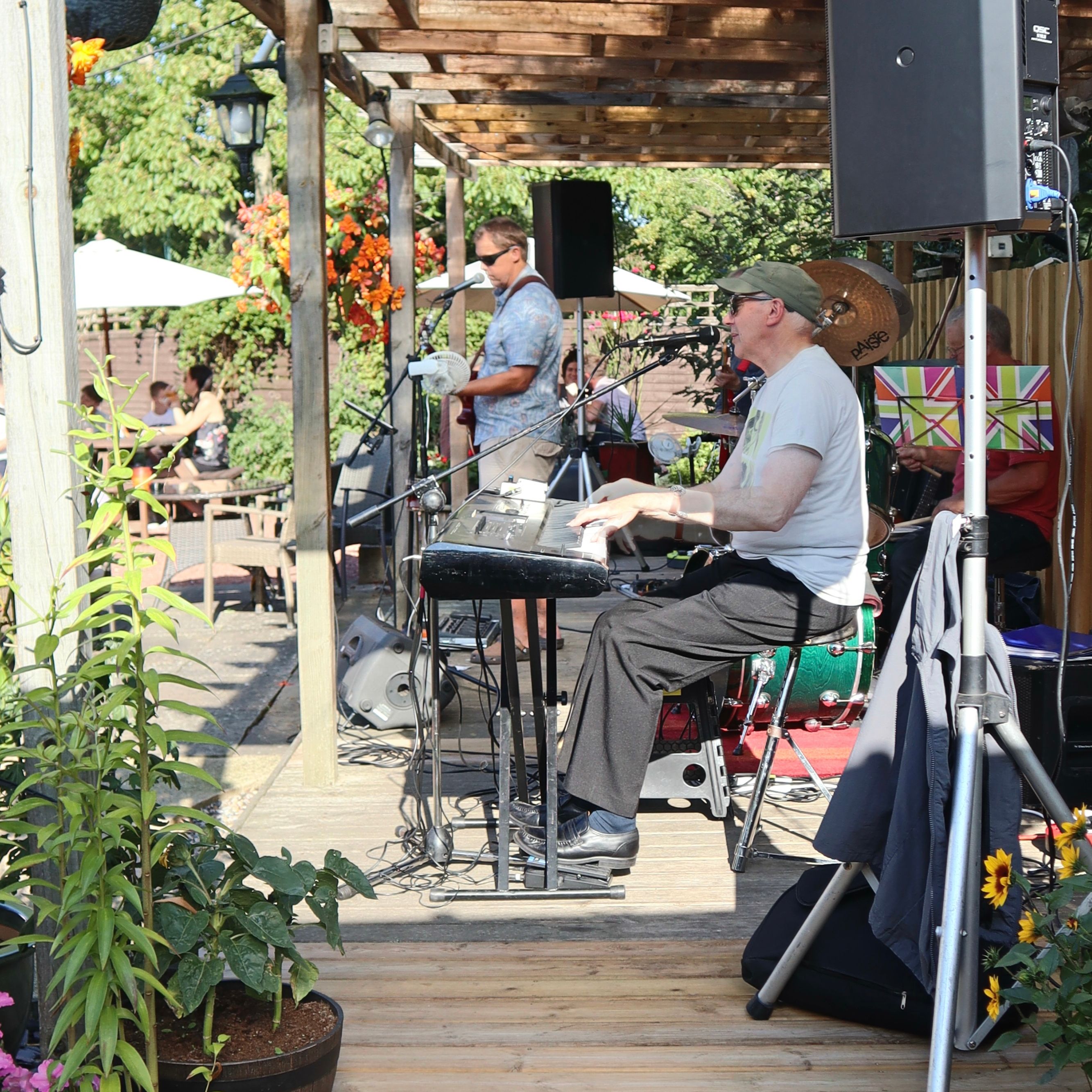 Outdoor band performance with a keyboard player and other musicians on a wooden stage surrounded by flowers and plants.